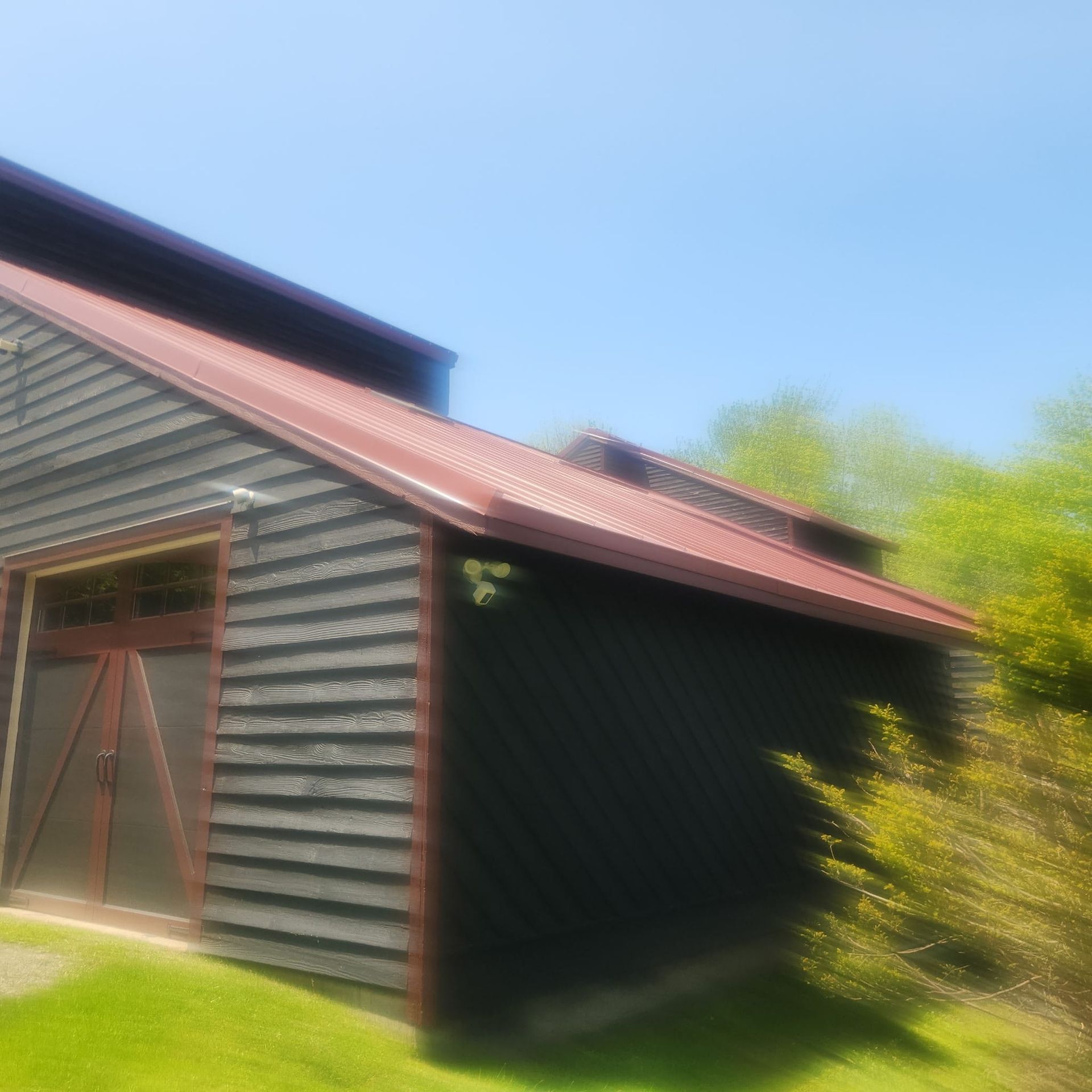 Red barn with brown doors and a sloped roof in a sunny yard, framed by green trees.