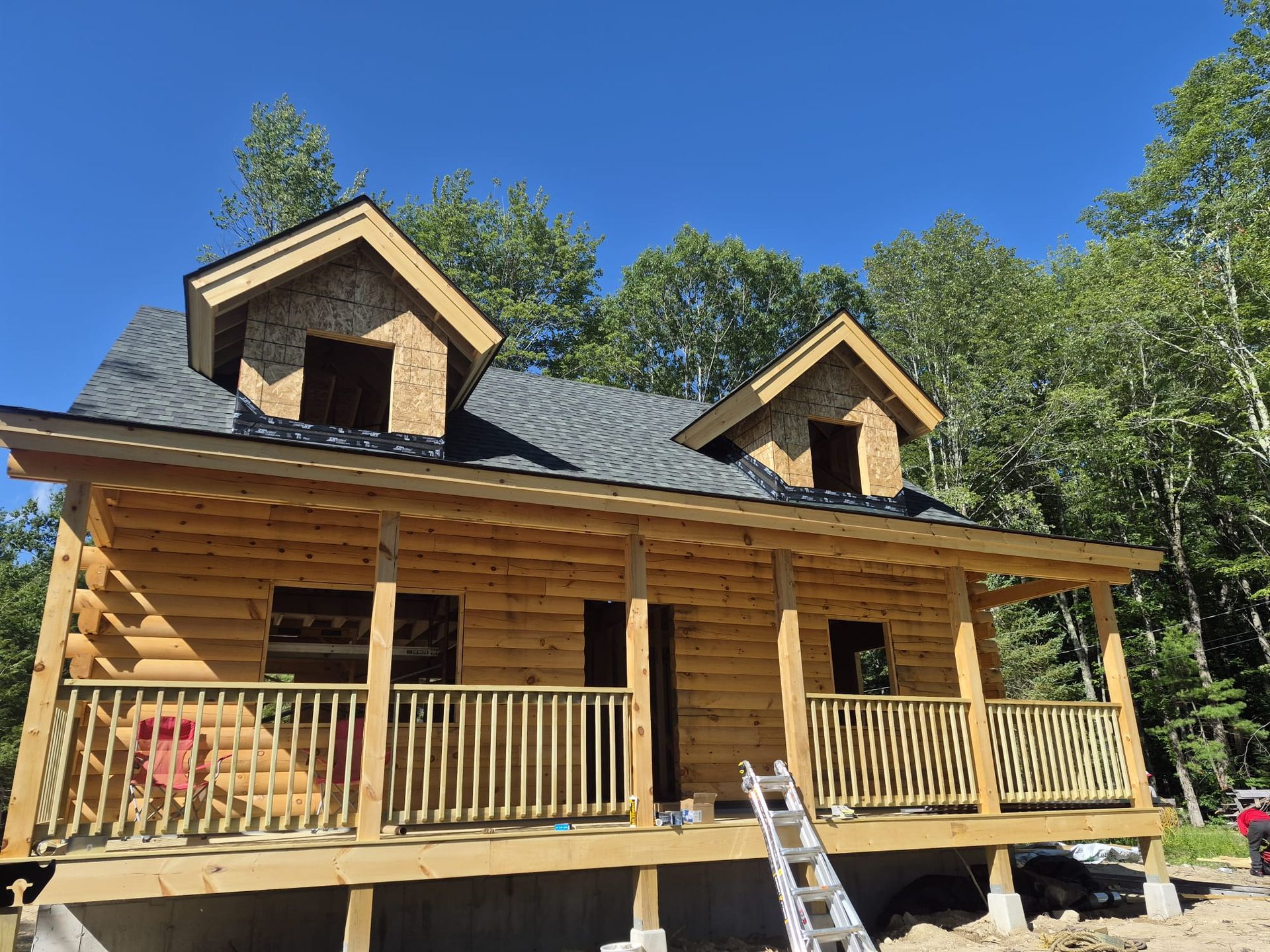 Wooden cabin under construction with a porch, ladder, and dormer windows in a sunny wooded area