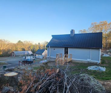 Rural house with dark roof, white siding, and a deck, surrounded by yard debris under a clear blue sky