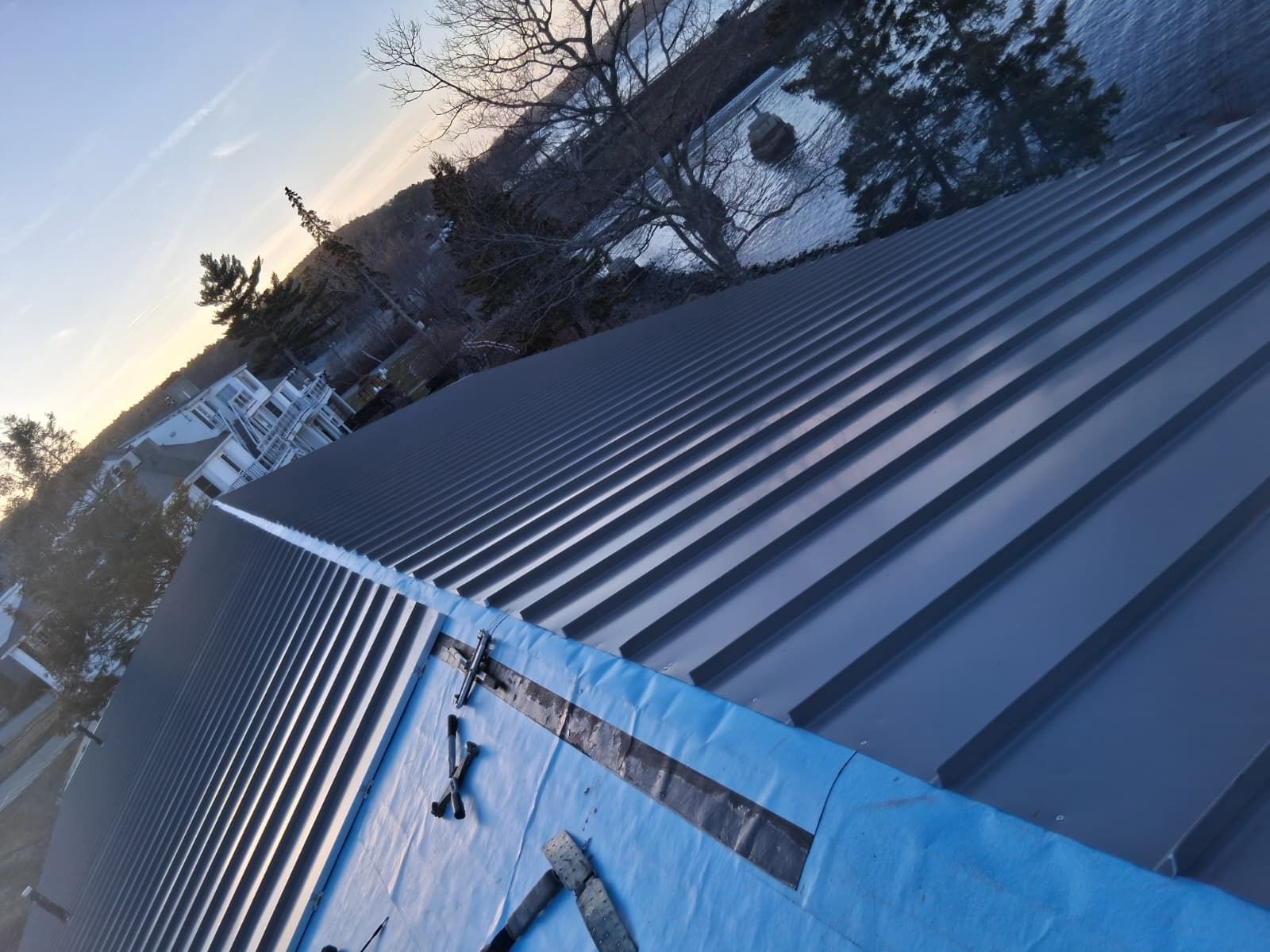 Snow-covered metal roof at sunset with a ladder leaning against it and trees in the background