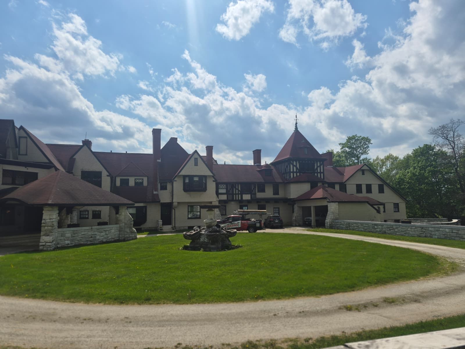 Large Tudor-style manor with red roofs and stone walls under a blue, cloudy sky, viewed across a green lawn and driveway