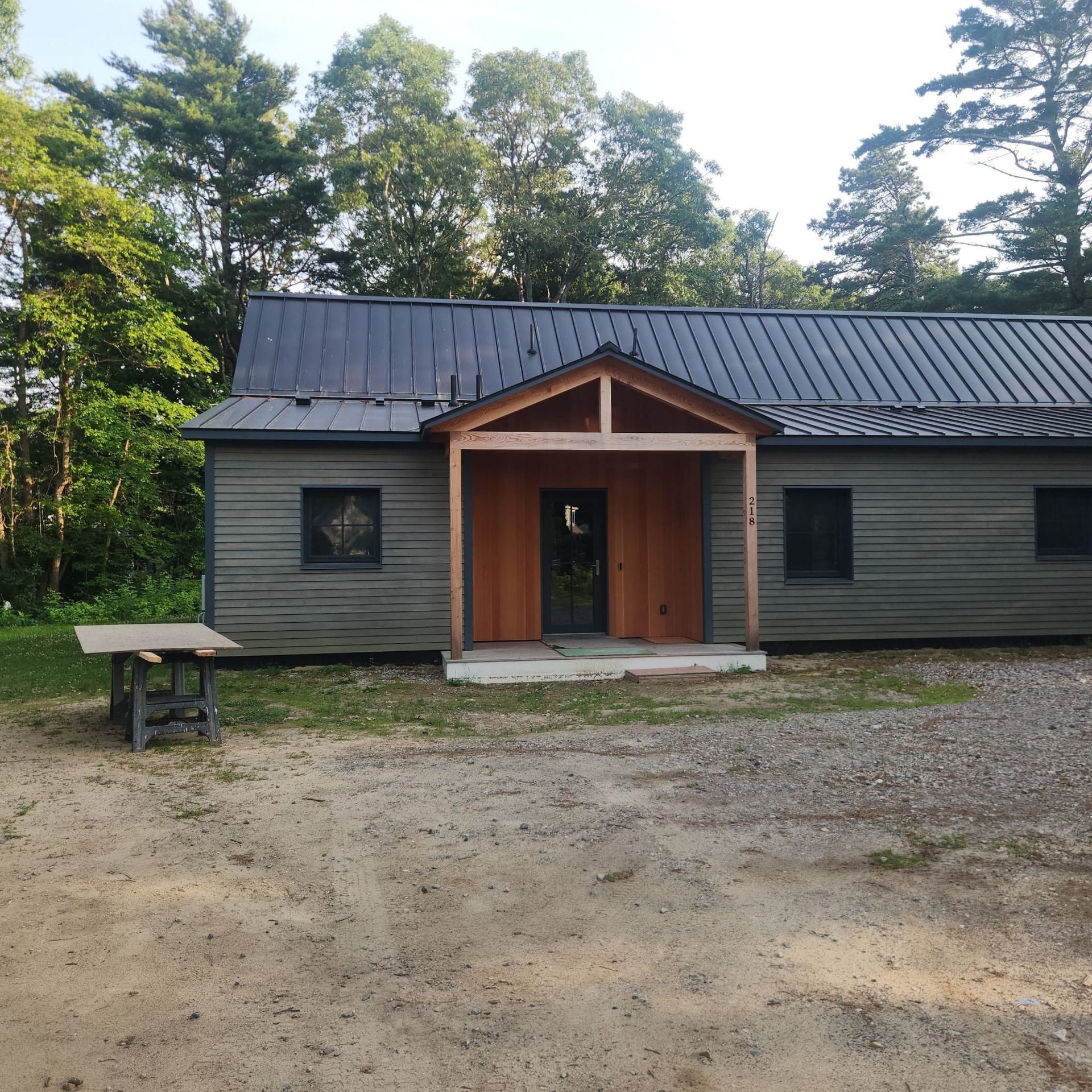 Small dark-gray cabin with a metal roof and wooden entrance, set in a wooded clearing with a dirt foreground.