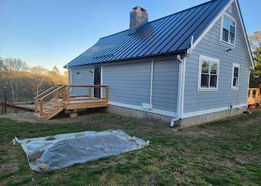 Blue-gray house with a metal roof and small wooden deck in a grassy yard at sunset