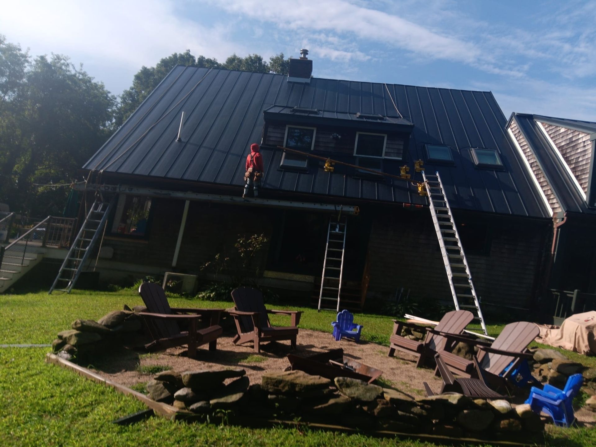 House with steep roof and deck under construction, ladders outside, fire pit and chairs in a grassy yard