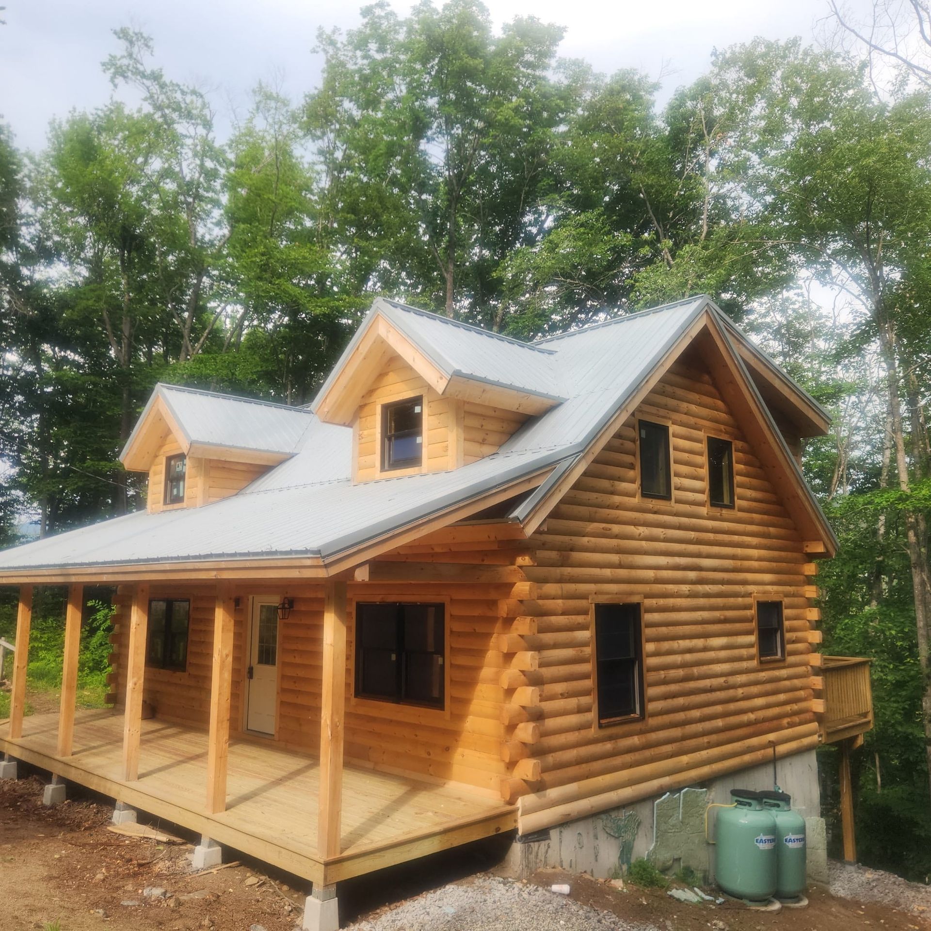 Log cabin-style house with wraparound porch, gray roof, and trees in the background