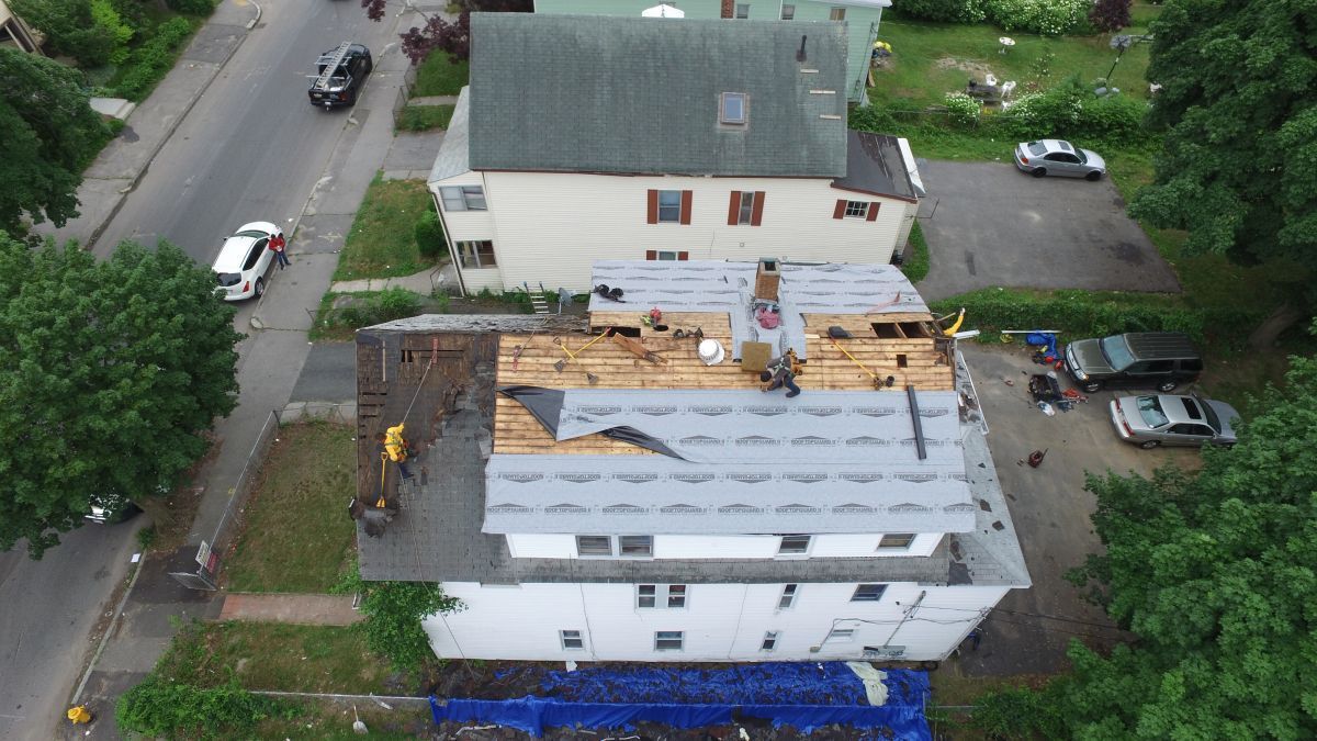 Aerial view of a house under construction with exposed roof framing beside a tree-lined street.