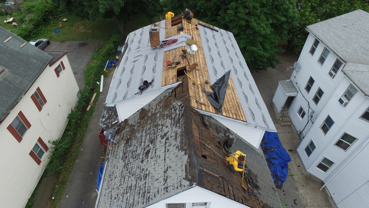 Aerial view of roof repair on a white building, with workers, exposed plywood, and blue tarp.