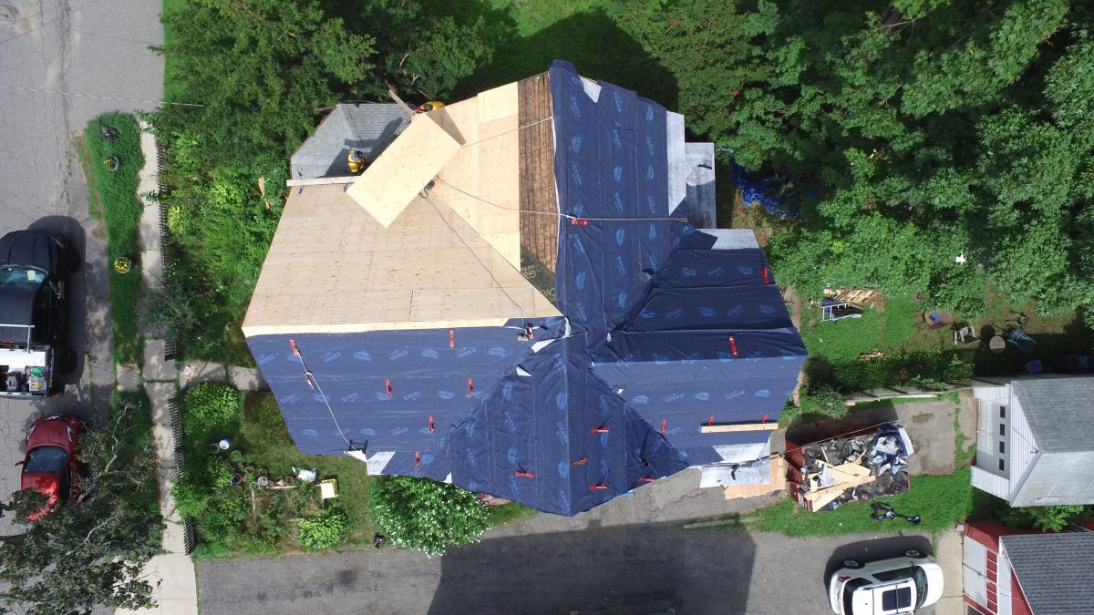 Aerial view of a house roof under construction with blue tarps and a yellow panel, surrounded by trees and parked cars