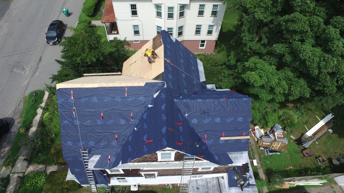 Aerial view of a house with a blue tarp-covered roof under repair, surrounded by trees and neighboring buildings.