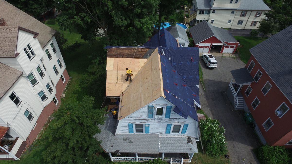Aerial view of a blue house with a partially replaced roof and nearby buildings and trees.