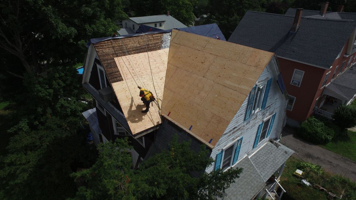 Roof being repaired on a house, with plywood sheathing exposed and a worker on the roof.