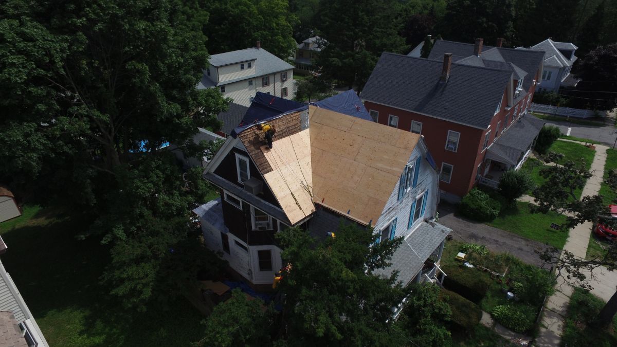 Aerial view of a house with a partially stripped roof beside a red brick home in a wooded neighborhood.
