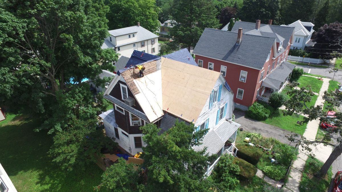 Aerial view of a house with a partially stripped roof beside a red home and green trees