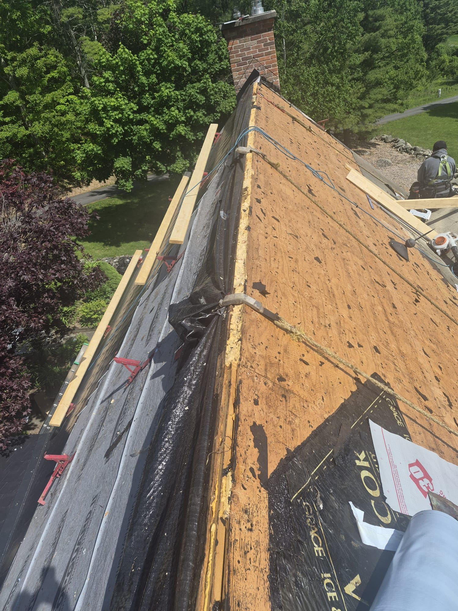 Roofer working on a steep roof with orange underlayment and safety gear beside trees and a brick chimney