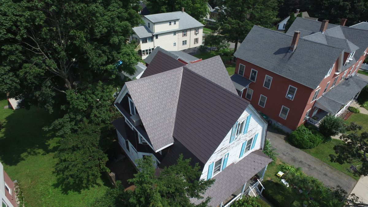 Aerial view of a gray-roofed house with blue trim beside a red house and trees.