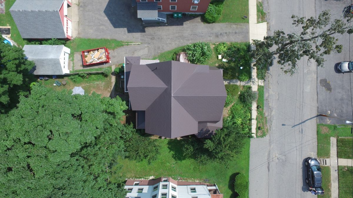 Aerial view of a suburban house with driveway, trees, and parked vehicles beside a road.
