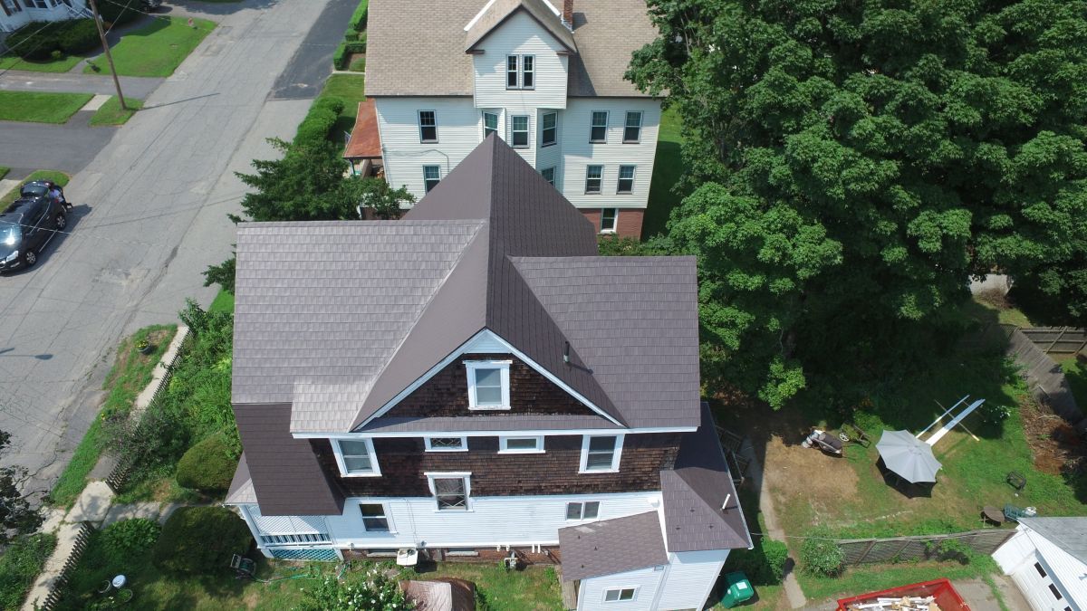 Aerial view of a brown house with a steep roof and a large tree beside it on a residential street