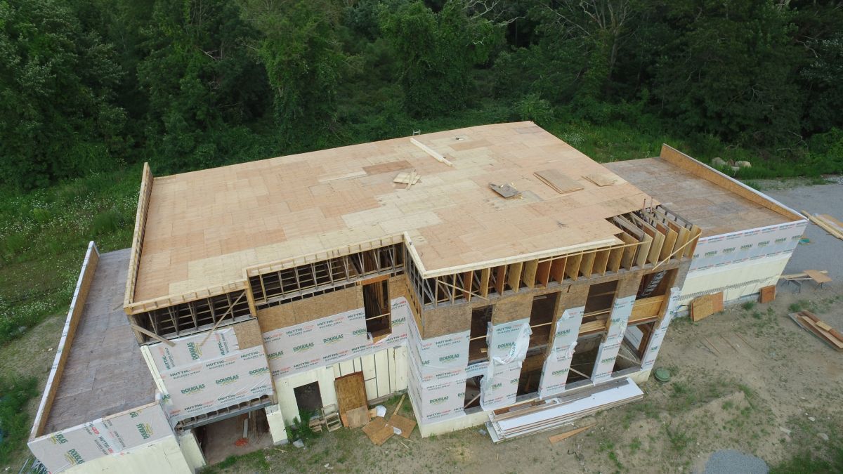 Aerial view of an unfinished multi-story building with exposed wooden roof framing beside a forest.