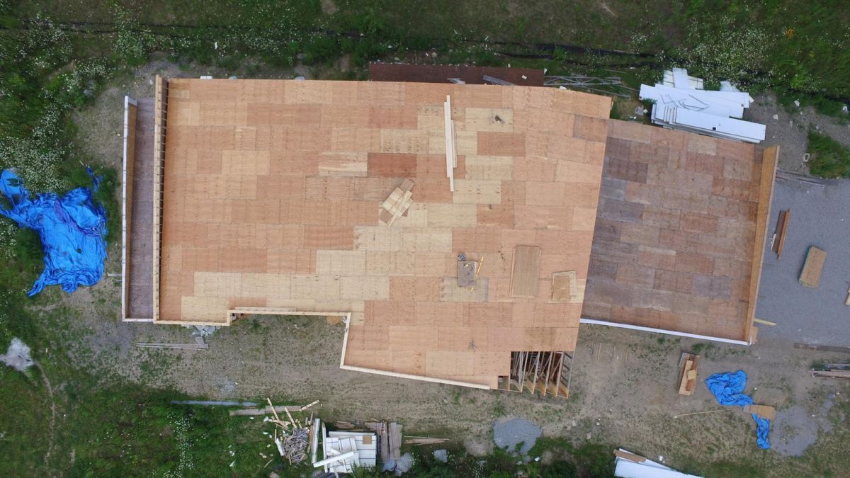 Top-down view of a house roof under construction with plywood sheathing and surrounding grass.