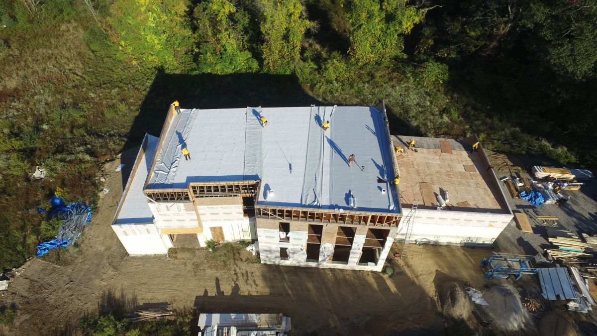 Aerial view of a large building with a white roof, surrounded by trees and construction materials.