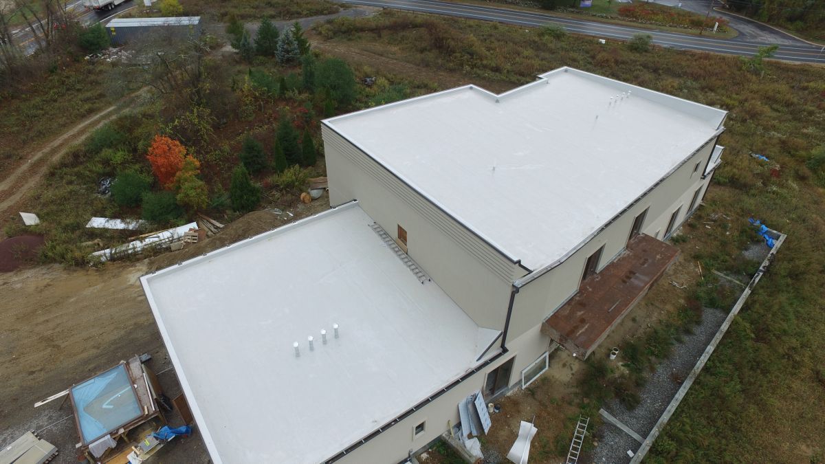 Aerial view of a beige building with a white roof beside trees and a dirt road
