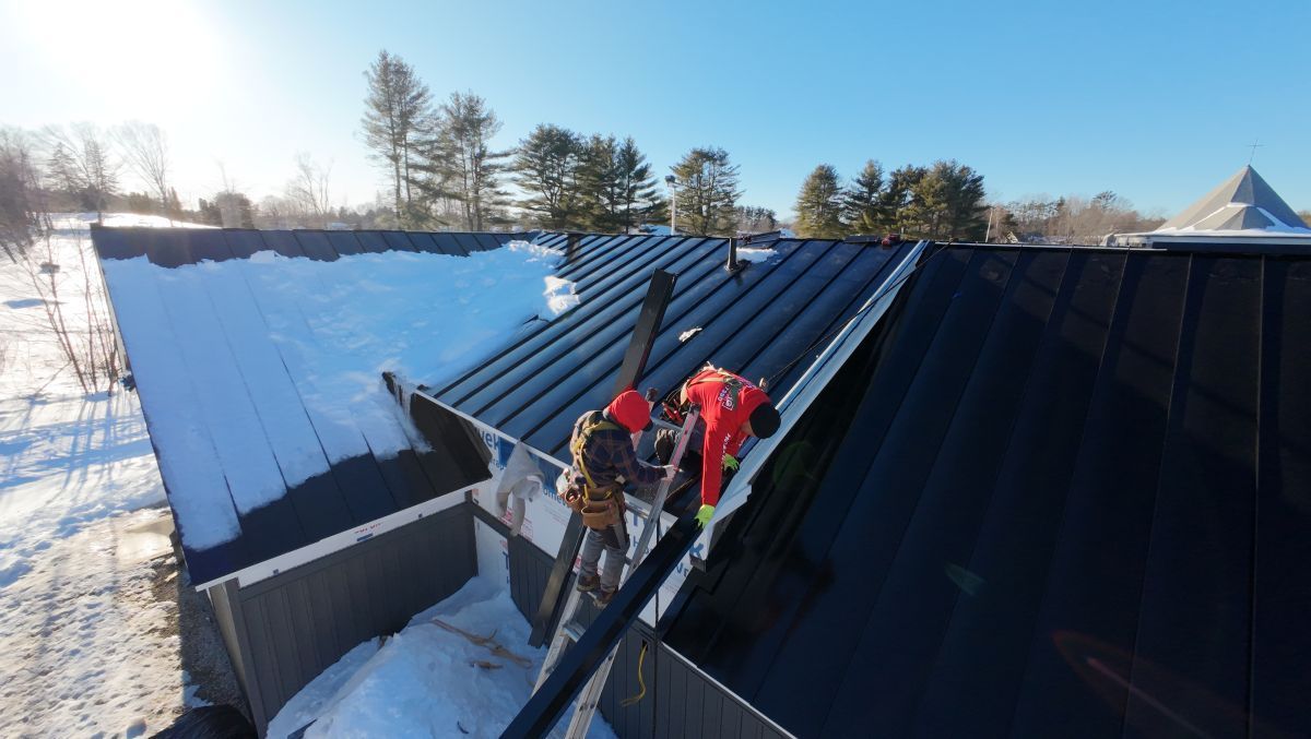 Workers in red jackets on a snowy black metal roof during maintenance