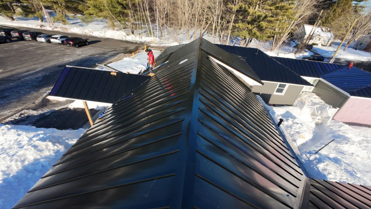 Snowy rooftop ridge with dark metal shingles and neighboring houses below on a sunny day