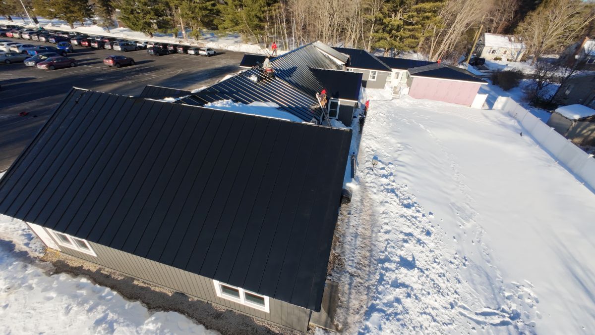 Aerial view of snow-covered rooftops beside a dark metal-roof building in a wooded area