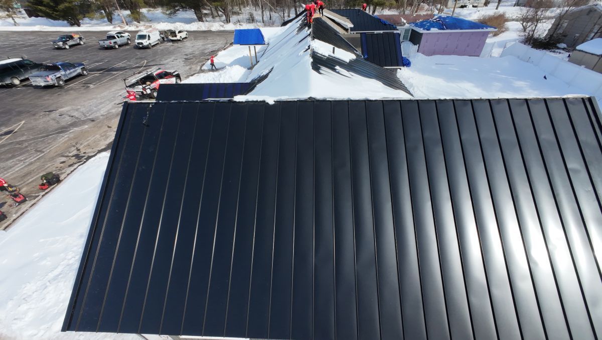 Snow-covered black metal roof under bright sunlight, with a parking lot and buildings in the background
