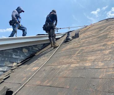 Two workers on a steep roof near the ridge, with blue sky above and shingles being repaired