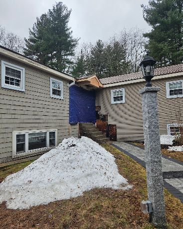 Snow pile outside beige house with blue tarp-covered entry and stone lamp post in front yard