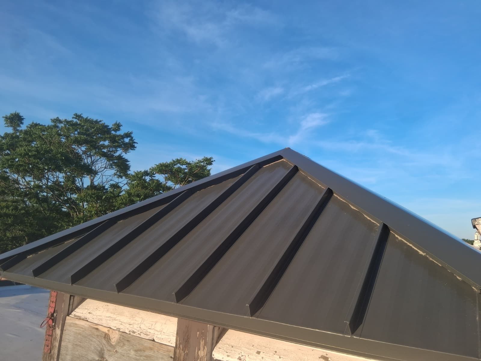 Dark gray metal roof on a small building under a blue sky with trees in the background