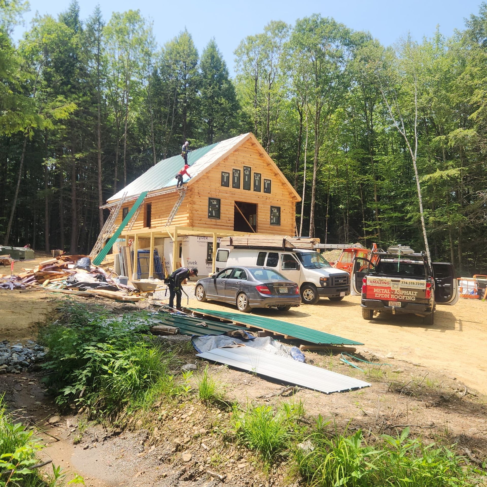 Wooden house under construction in a forest clearing with trucks, cars, and building materials outside.