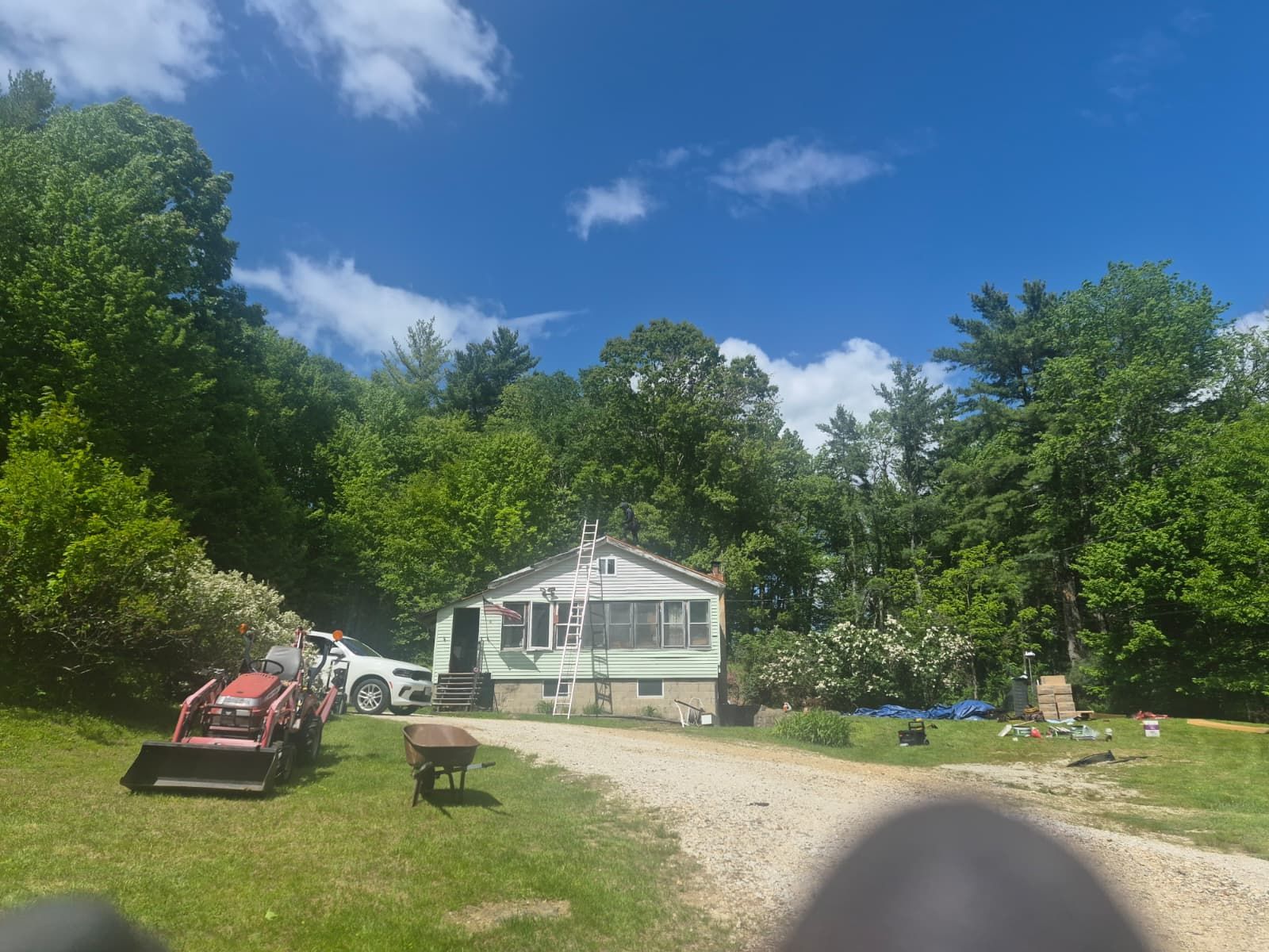 Rural white house with porch and gravel driveway, surrounded by trees under a bright blue sky.