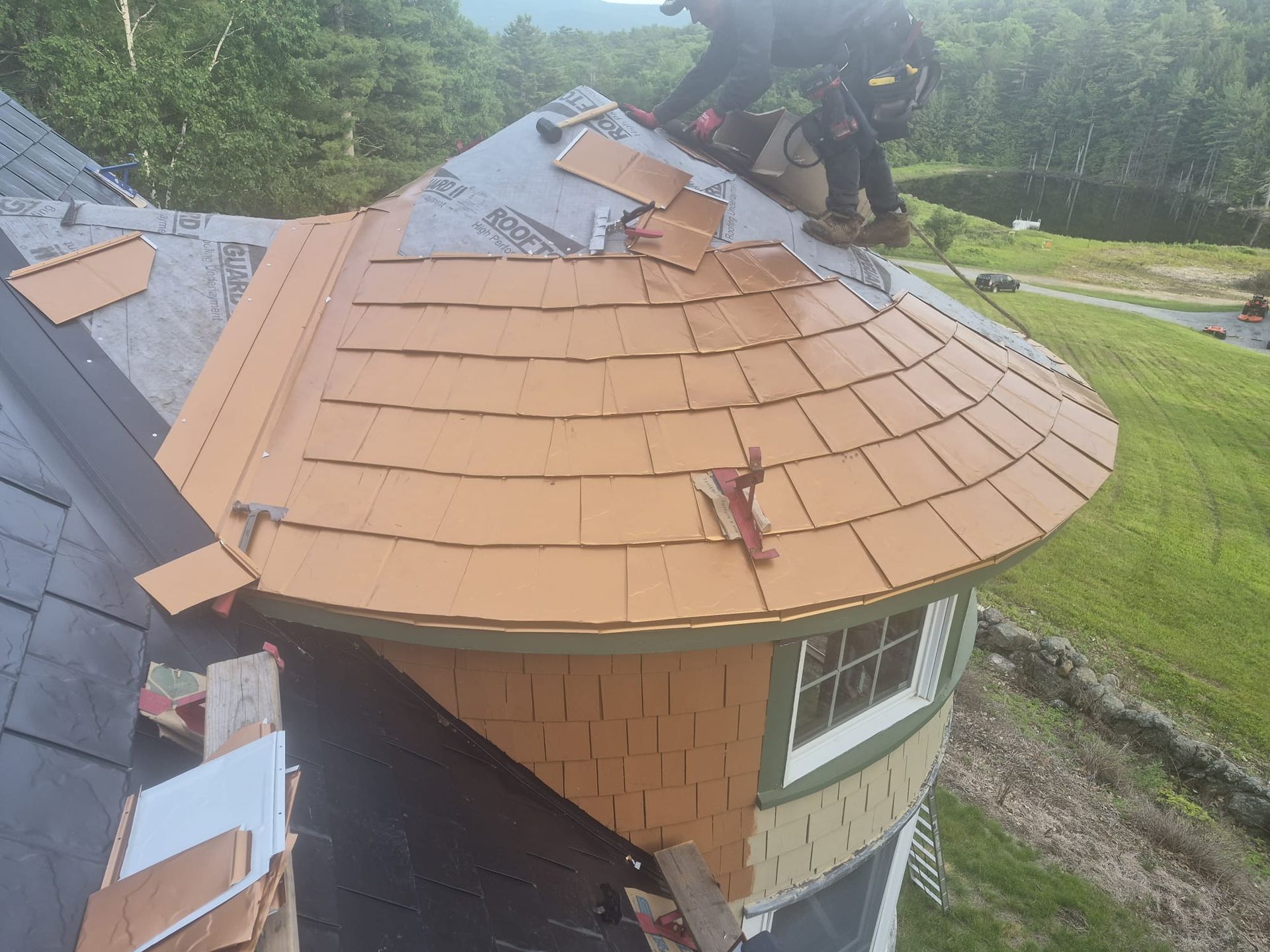 Roofers installing orange shingles on a round house roof with a ladder and tools nearby.
