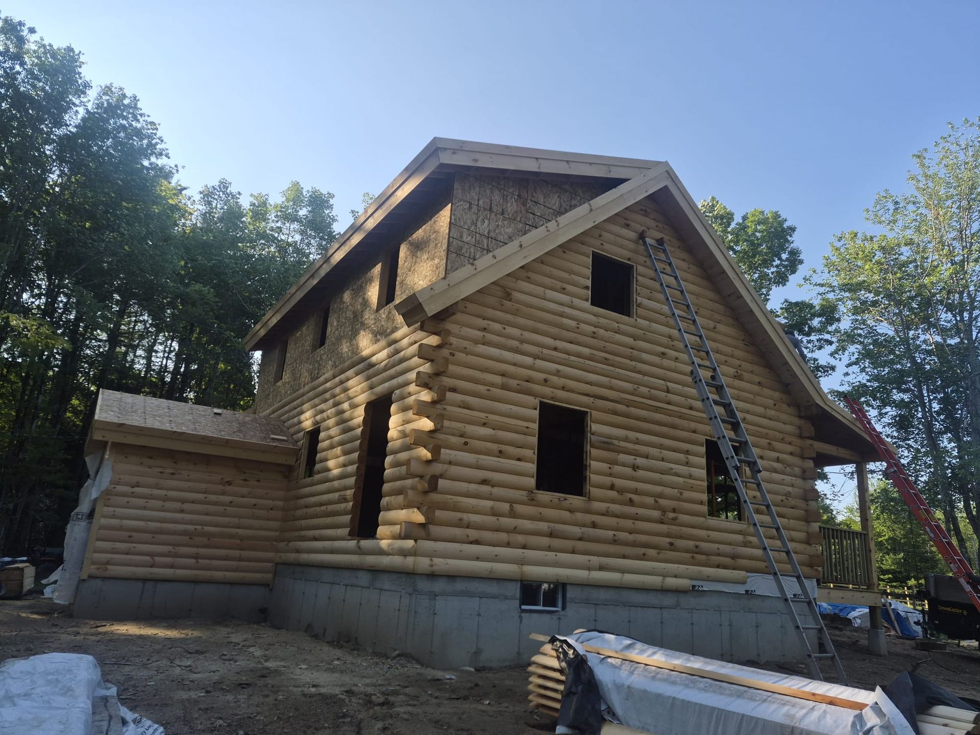 Log cabin under construction with ladder, concrete foundation, and trees in the background