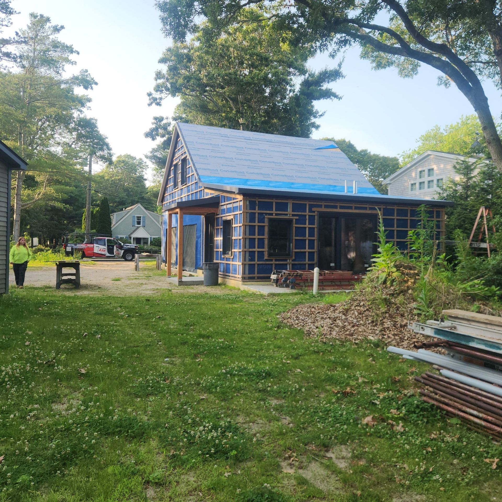 Blue-roofed shed under construction in a grassy yard with trees and stacked boards