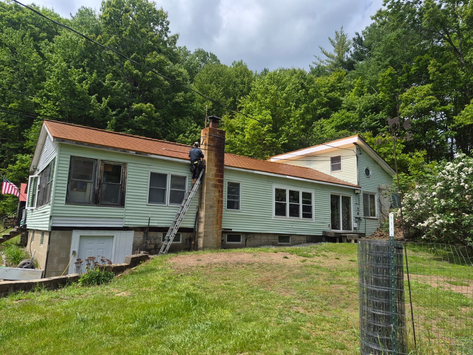 Mint-green house with damaged siding and brick chimney on a grassy hillside, surrounded by trees