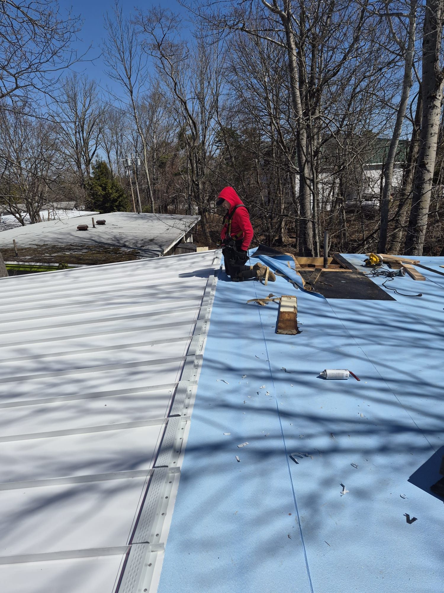 Worker in red jacket on a snowy roof, installing light blue roofing panels under bare trees.