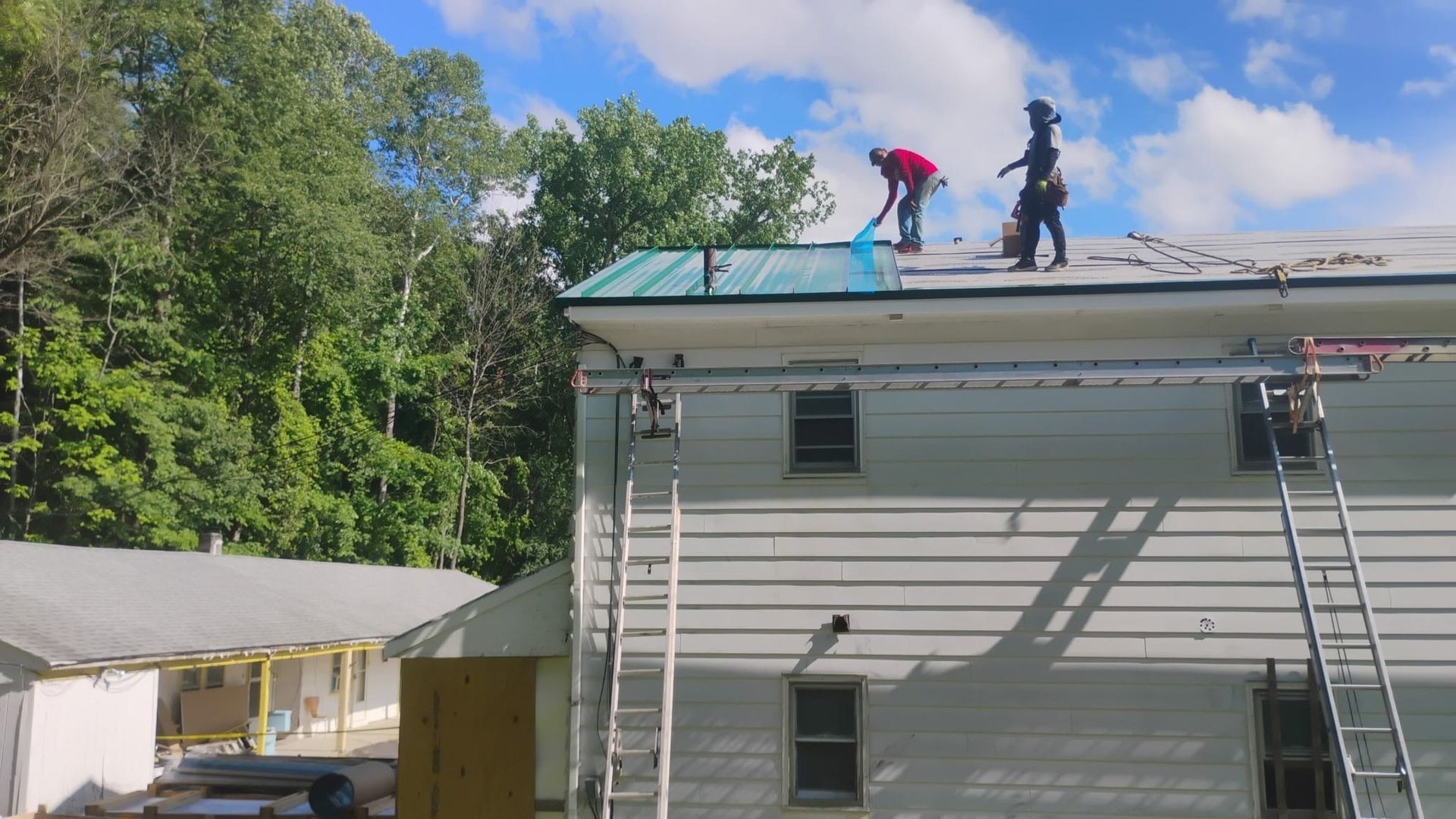 Two workers on a roof, one carrying a red bucket, beside a white house under a blue sky.