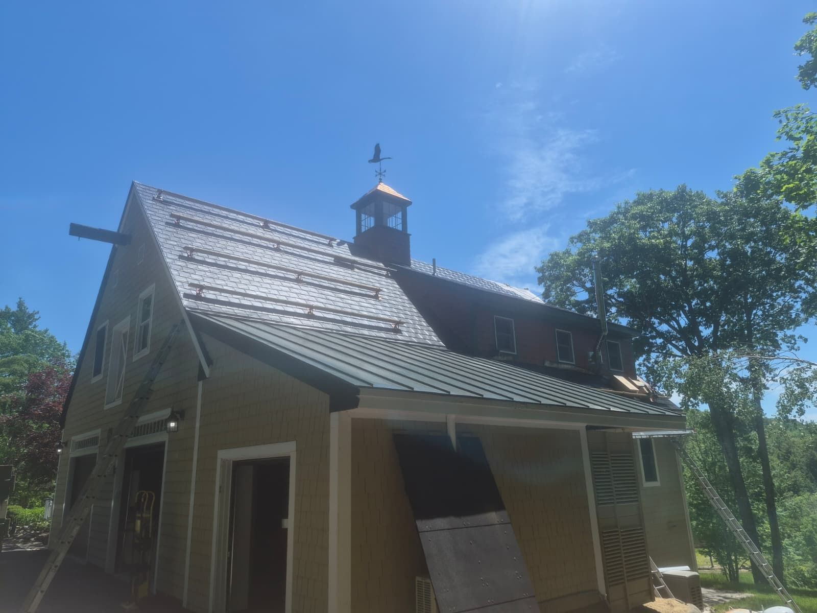 Small beige house with a gray roof and cupola under a bright blue sky, surrounded by trees