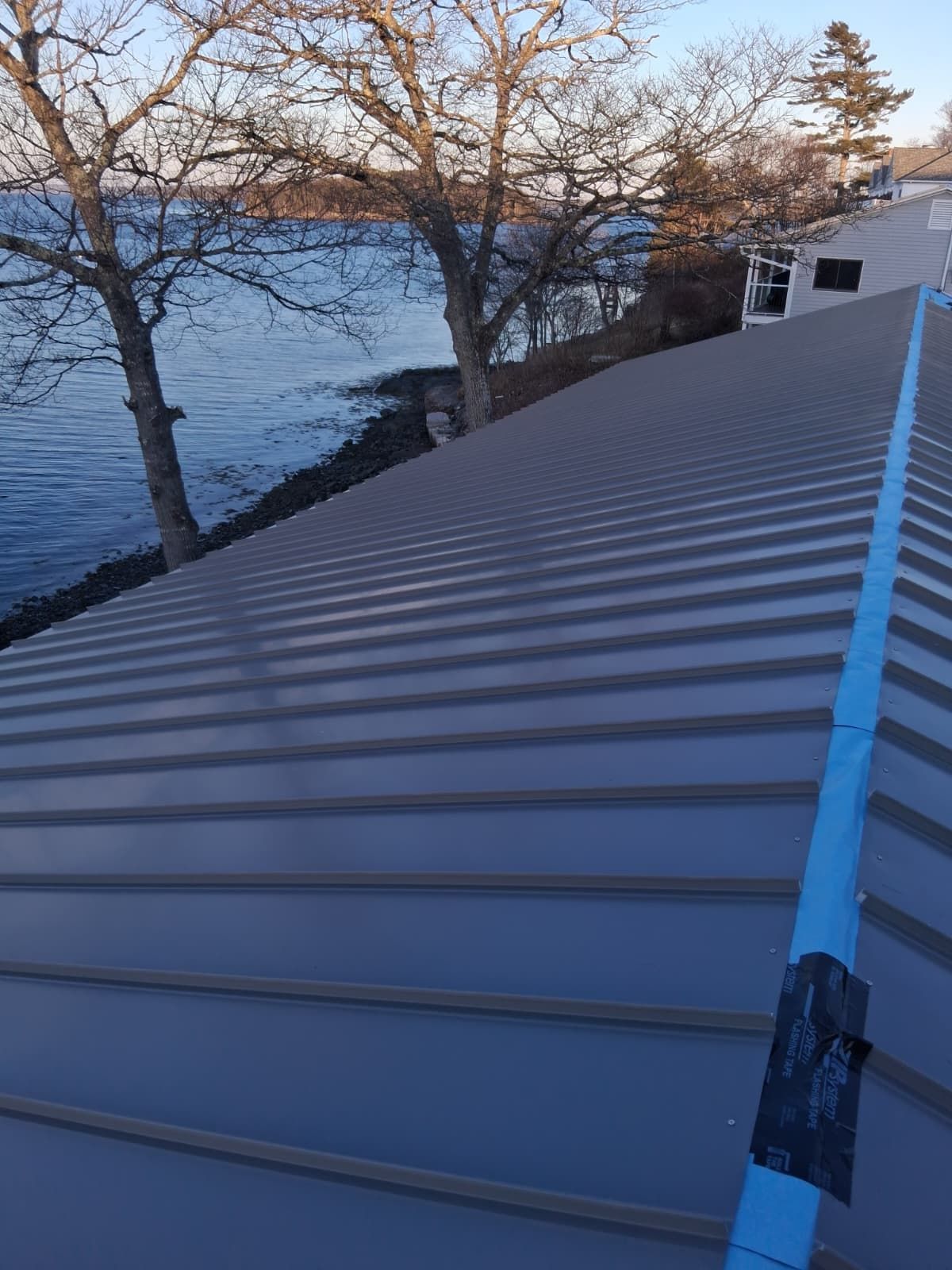 Corrugated metal rooftop by a lake at sunset, with trees and a blue seam line running up the roof.