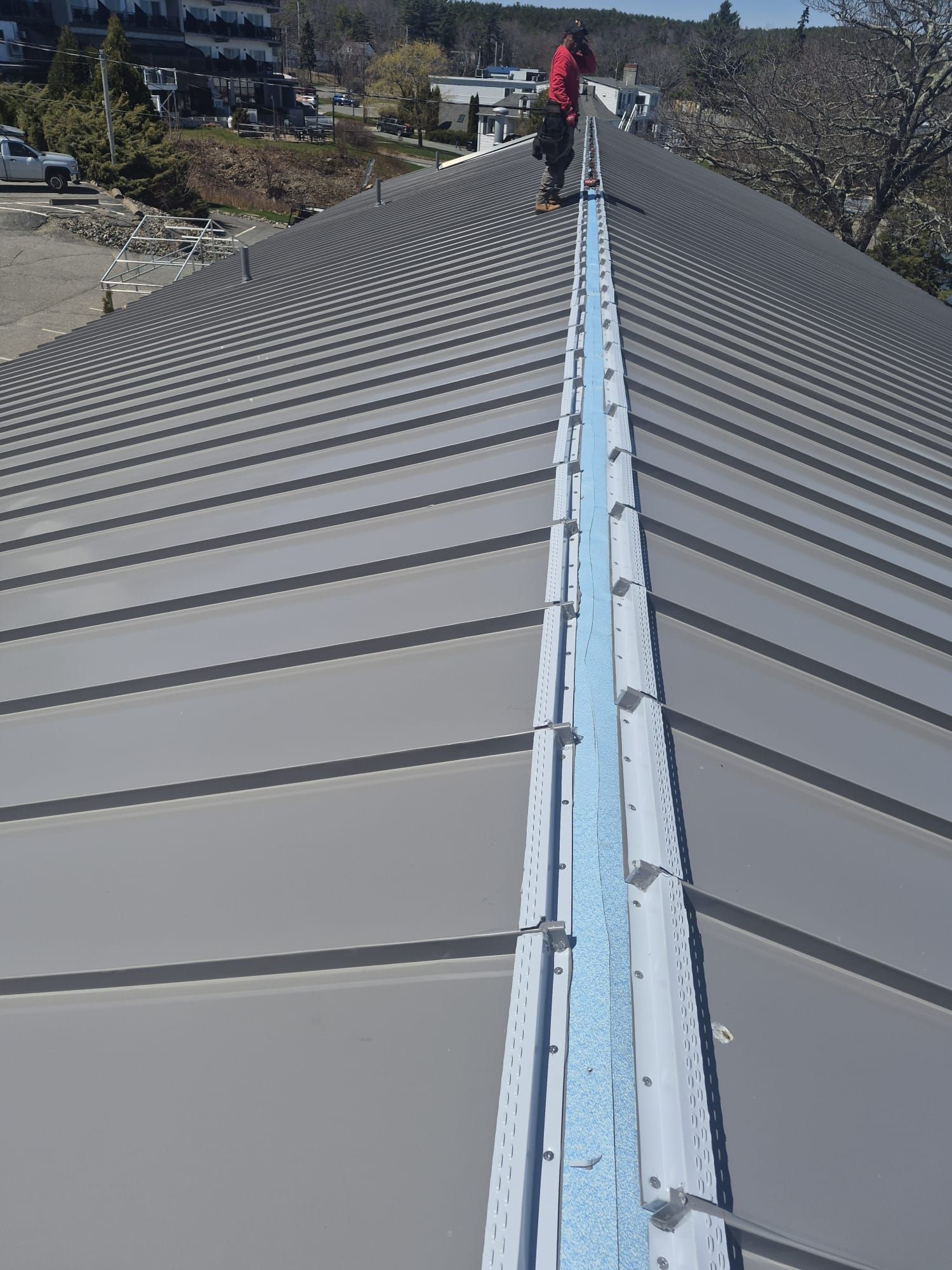 Person inspecting a long gray metal roof with a central ridge under a clear sky.