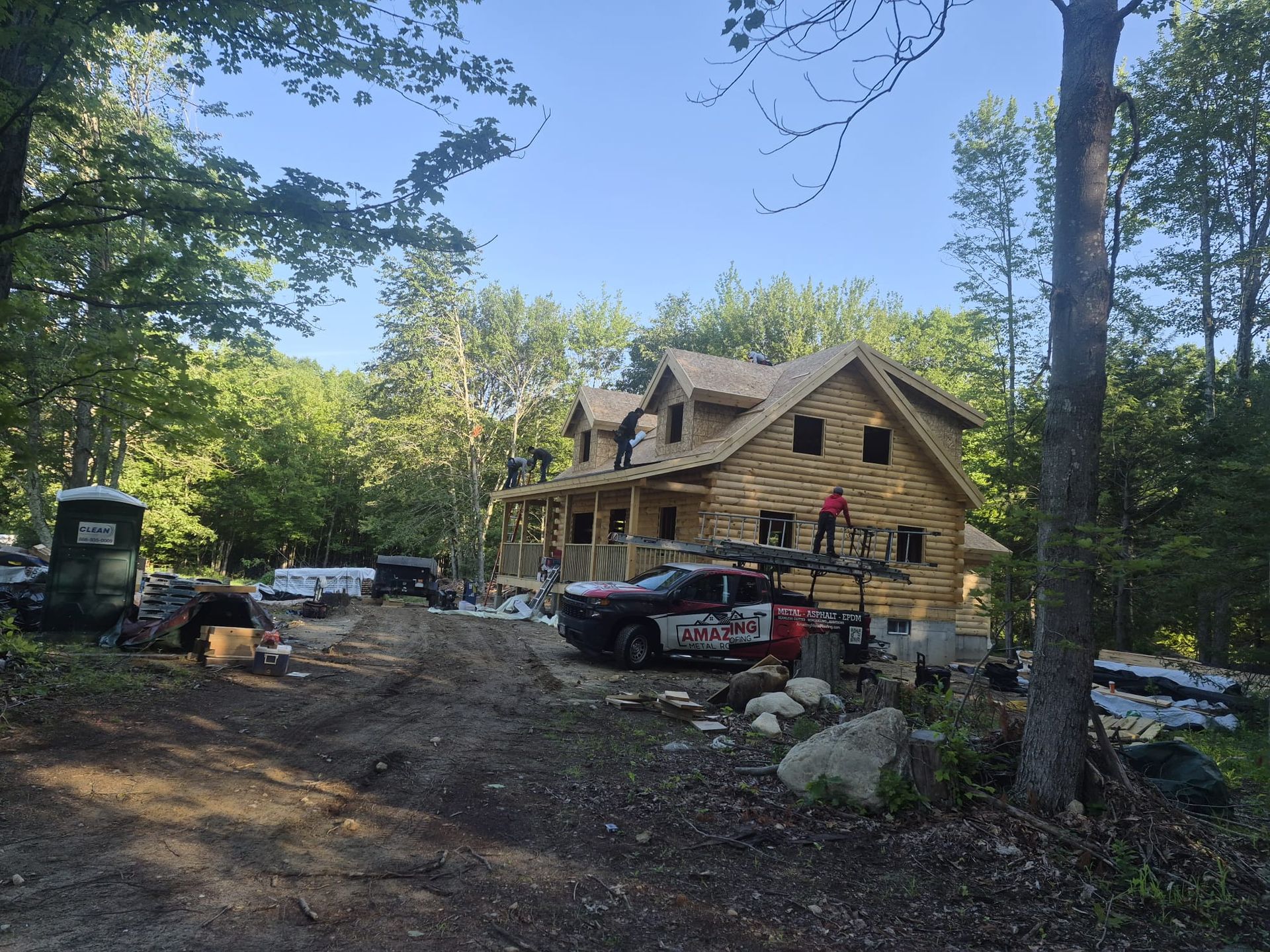 Log cabin under construction in a wooded clearing, with workers, parked vehicles, and building materials.