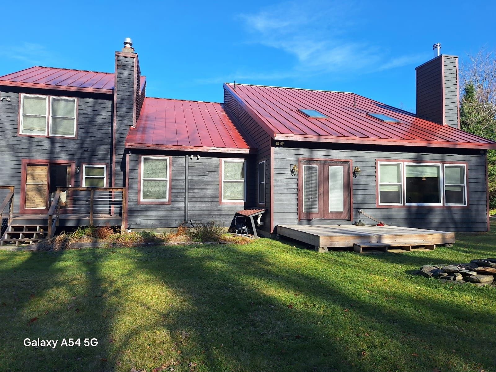 Gray two-story house with red metal roof, stone chimney, and large windows in a sunny yard
