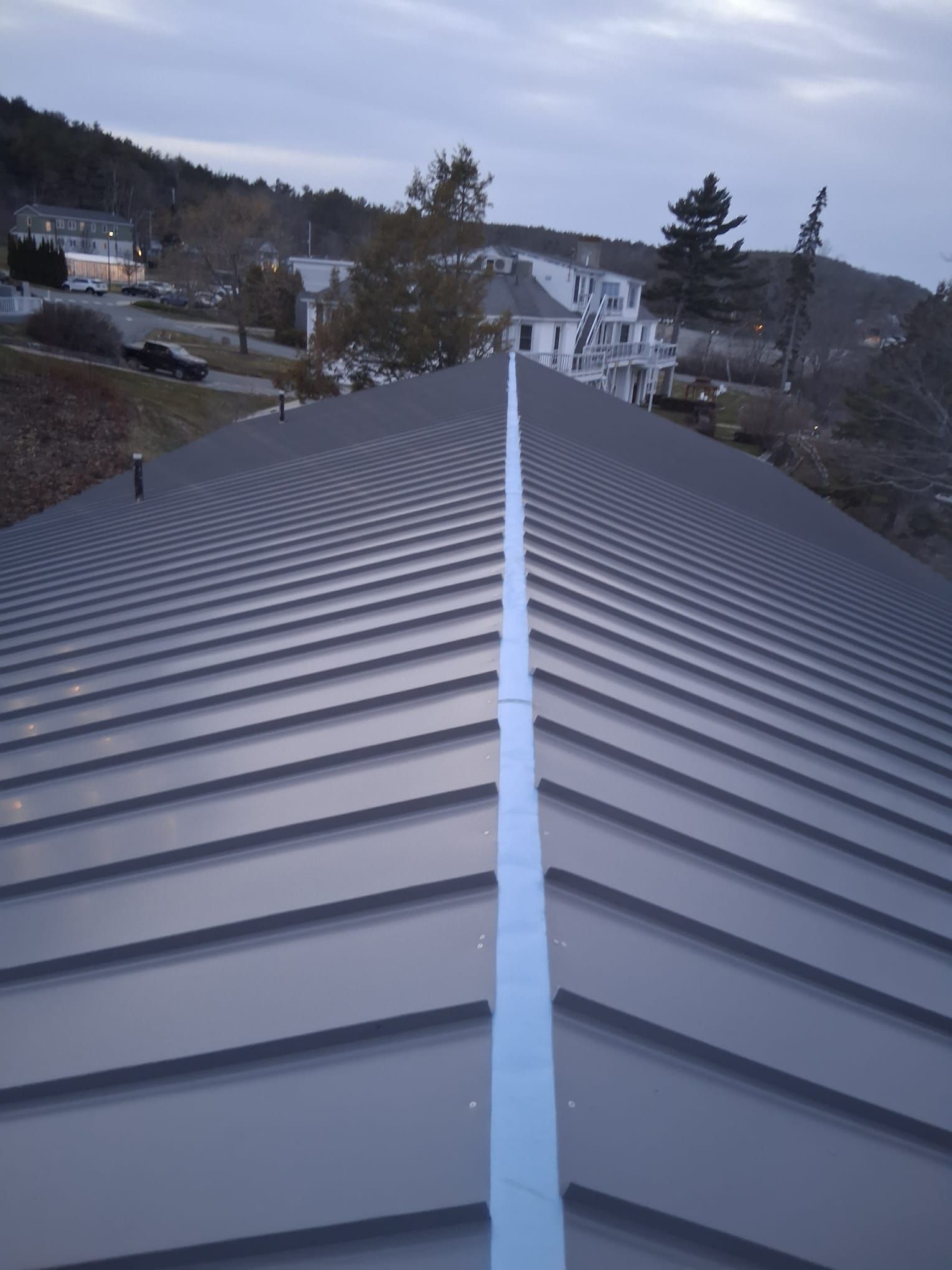 Corrugated metal roof ridge leading toward houses and trees under a cloudy sky