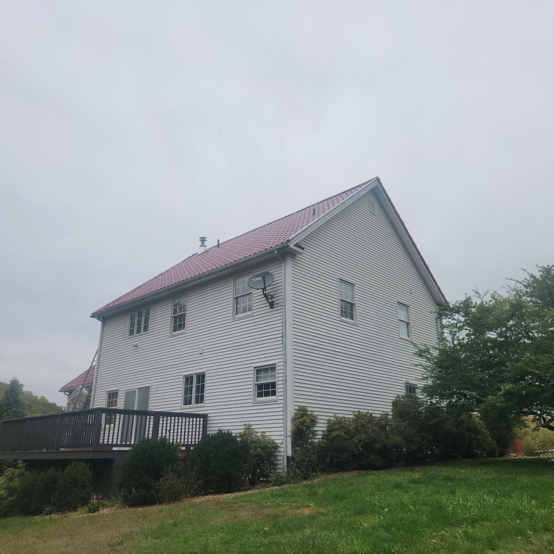White farmhouse with red roof on a grassy hill under a gray sky, with a porch and trees nearby