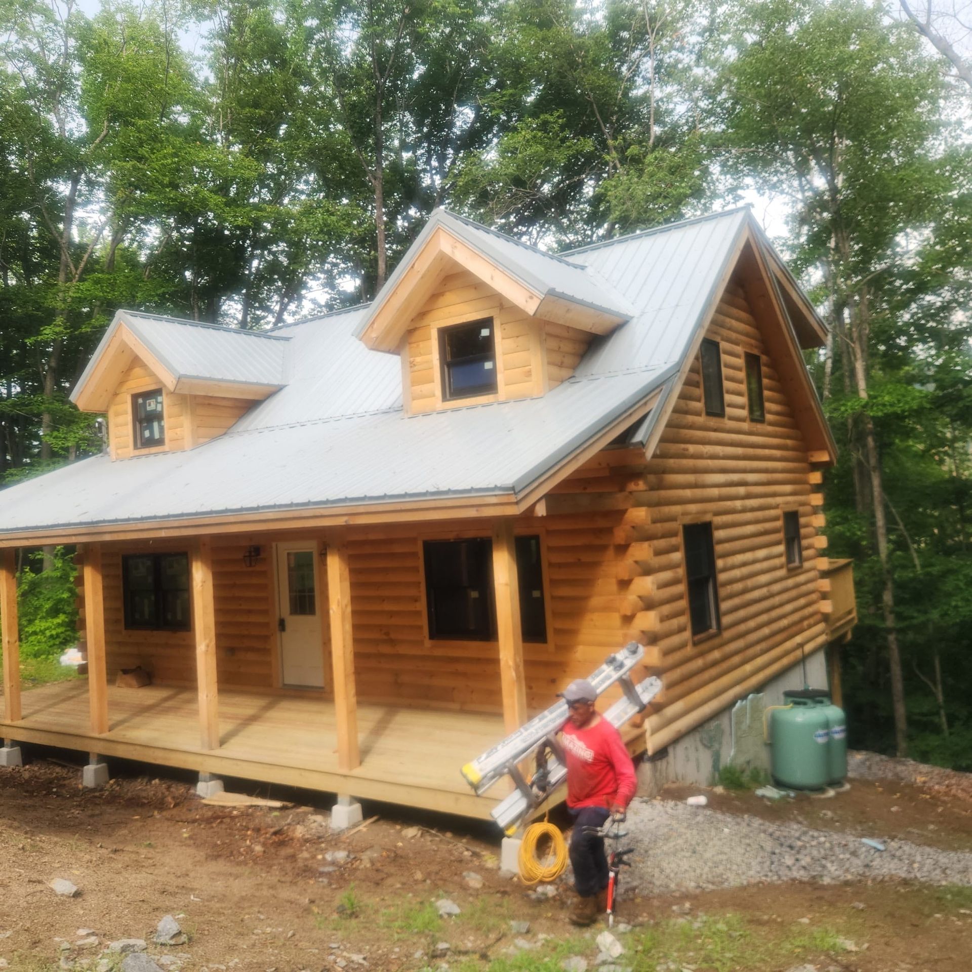 Log cabin under construction with porch; worker carrying a ladder in front of trees