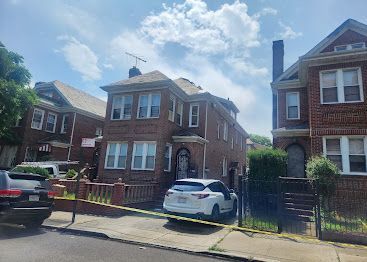 Brick houses on a sunny street with parked cars and a black iron fence
