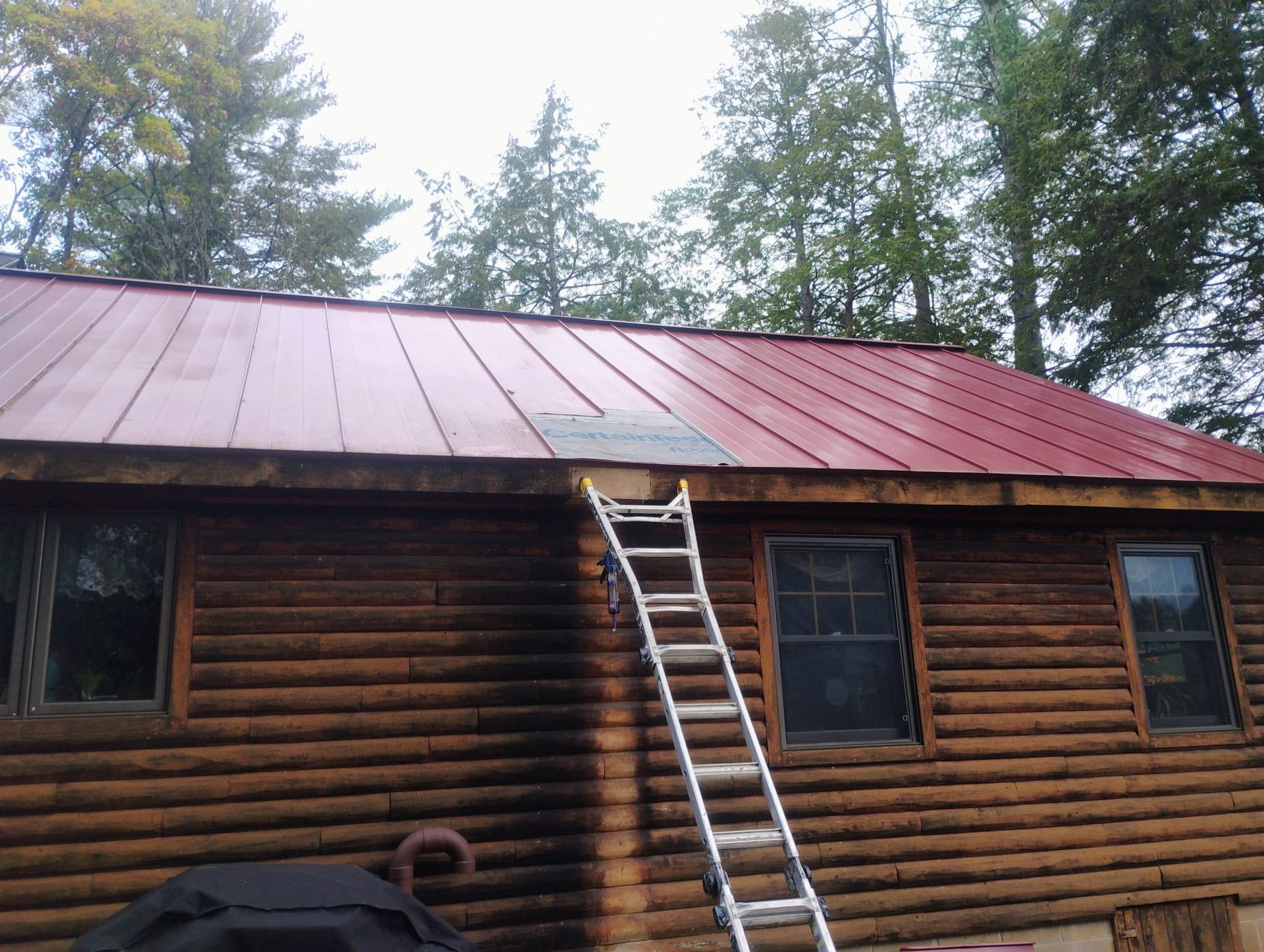 Ladder leaning on log cabin roof with red and rusted metal panels amid trees
